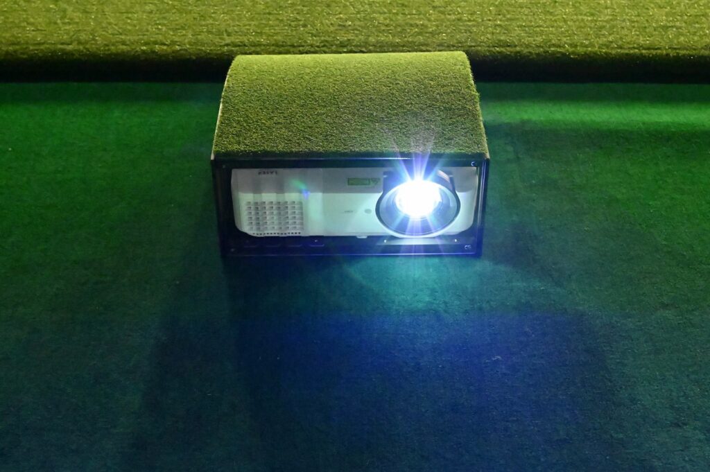 Carl's Place floor mounted projector enclosure with a turf top on a green mat and the projector lens lit
