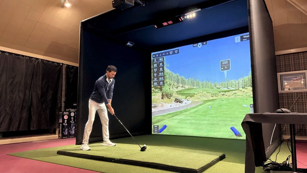Jay addressing a driver on a raised hitting platform in a SIG PRO enclosure under an Uneekor EYE XO at Tyrone Hills Golf Club