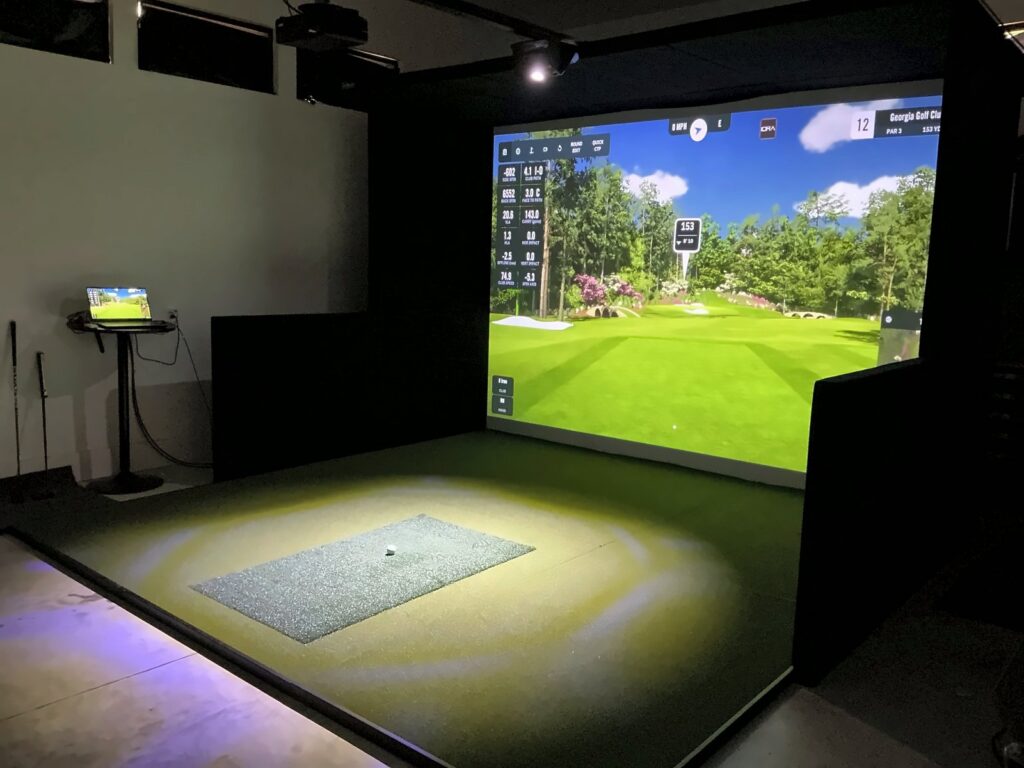 A hitting mat set flush into the turf floor of a GolfIN SimBooth 2 golf simulator, lit by a spotlight