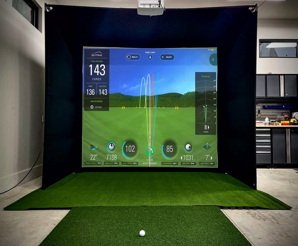 A garage golf simulator with a black enclosure, turf, a SkyTrak screen showing shot data, and a projector hung from the top of the frame