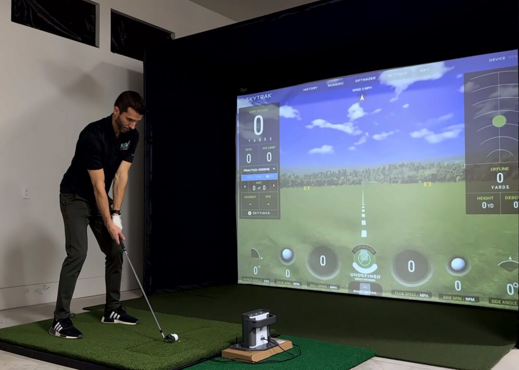 Jay addressing an iron with a SkyTrak beside the ball and the SkyTrak range on the screen of a Carl's Place DIY enclosure